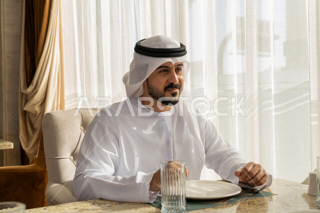 قضاء الأوقات الممتعة في الخارج, إيماءات السعادة و السرور, رجل عربي خليجي إماراتي مبتسم يرتدي الكندورة و الغترة البيضاء يجلس في إحدى المطاعم في الإمارات, أنشطة ترفيهية مسلية, الاستمتاع بالأجواء المميزة في المطعم
