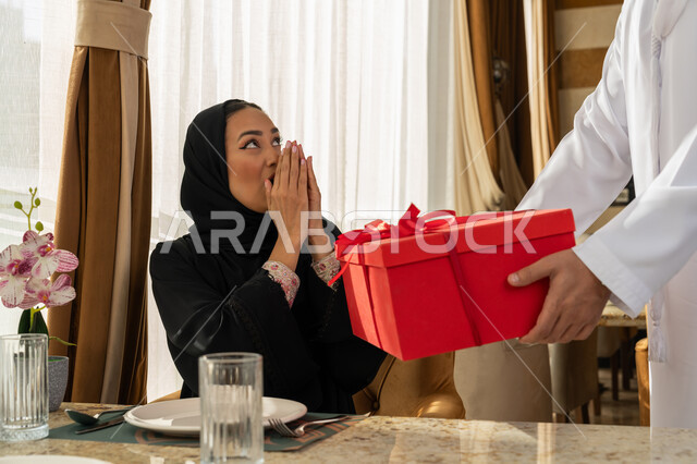 تعابير الاندهاش و الفرح, تقديم الهدايا بذكرى يوم الزواج، التعبير عن الألفة والمحبة بين الزوجين, قضاء أوقات ممتعة مع الأسرة, رجل عربي خليجي إماراتي يرتدي الكندورة البيضاء يقدم لزوجته هديه في علبة حمراء, الاحتفال بالمناسبات و الأعياد العائلية
