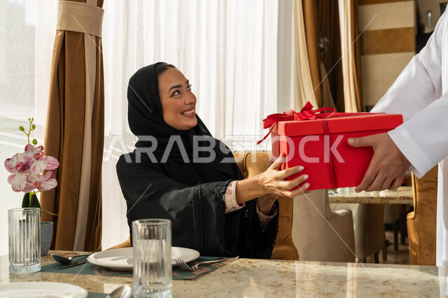 تقديم الهدايا بذكرى يوم الزواج، التعبير عن الألفة والمحبة بين الزوجين, قضاء أوقات ممتعة مع الأسرة, رجل عربي خليجي إماراتي يرتدي الكندورة البيضاء يقدم لزوجته هديه في علبة حمراء, الاحتفال بالمناسبات و الأعياد العائلية