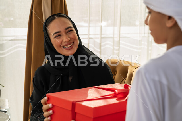 الاحتفال بمناسبة عيد الأم, إيماءات الفرح و البهجة, مفهوم احترام وتقدير وبر الوالدين, إدخال السعادة والسرور إلى قلب الأهل, ولد عربي خليجي إماراتي يرتدي الكندورة و العمامة البيضاء يقوم بتقديم علبة هدايا حمراء لوالدته