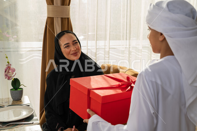 مفهوم احترام وتقدير وبر الوالدين, إدخال السعادة والسرور إلى قلب الأهل, ولد عربي خليجي إماراتي يرتدي الكندورة و العمامة البيضاء يقوم بتقديم علبة هدايا حمراء لوالدته, إيماءات الفرح و البهجة, تقديم الهدايا بمناسبة عيد الأم