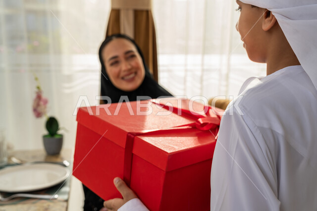 إدخال السعادة والسرور إلى قلب الأهل، مفهوم احترام وتقدير وبر الوالدين, ولد عربي خليجي إماراتي يرتدي الكندورة و العمامة البيضاء يقوم بتقديم علبة هدايا حمراء لوالدته, إيماءات الفرح و البهجة, تقديم الهدايا بمناسبة عيد الأم