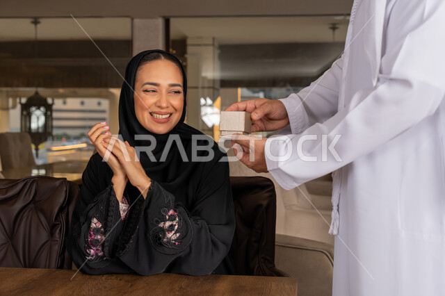 الأجواء الرومنسية الرائعة في المطعم, قضاء الأوقات الممتعة مع الأسرة, الاستمتاع باللحظات العائلية المميزة, تقديم الهدايا بمناسبة ذكرى يوم الزواج, رجل عربي خليجي إماراتي يرتدي الكندورة البيضاء يقدم لزوجته هدية تعبيراً عن الالفة و المحبة