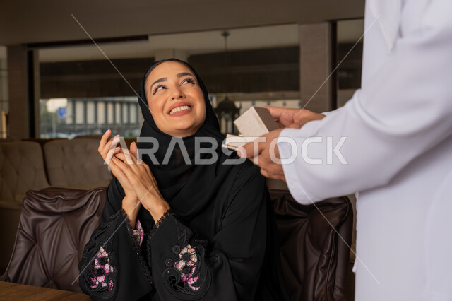 إيماءات السعادة و السرور, التعبير عن الألفة والمحبة بين الزوجين, تقديم الهدايا بمناسبة ذكرى يوم الزواج, قضاء الأوقات العائلية الممتعة, رجل عربي خليجي إماراتي يرتدي الكندورة البيضاء يقدم لزوجته هدية مميزة, الأجواء الرومنسية الرائعة في المطعم