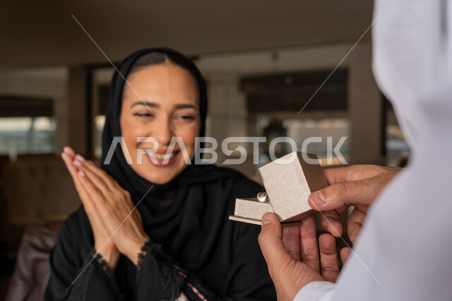 التعبير عن الألفة والمحبة بين الزوجين, تقديم الهدايا بمناسبة ذكرى يوم الزواج، إيماءات السعادة و السرور, قضاء الأوقات العائلية الممتعة, رجل عربي خليجي إماراتي يرتدي الكندورة البيضاء يقدم لزوجته هدية مميزة, الأجواء الرومنسية الرائعة في المطعم