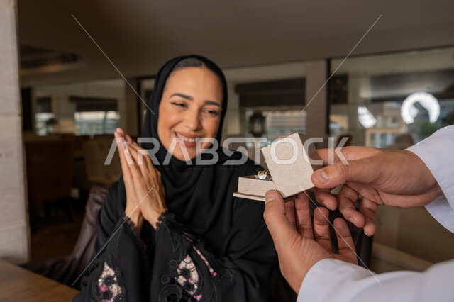 تقديم الهدايا بمناسبة ذكرى يوم الزواج، التعبير عن الألفة والمحبة بين الزوجين, إيماءات السعادة و السرور, قضاء الأوقات العائلية الممتعة, رجل عربي خليجي إماراتي يرتدي الكندورة البيضاء يقدم لزوجته هدية مميزة, الأجواء الرومنسية الرائعة في المطعم