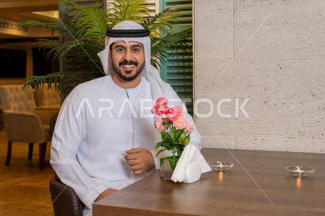 النظر إلى الكاميرا بإيماءات السعادة و السرور, الاستمتاع في أوقات الفراغ في إحدى المطاعم, قضاء الأوقات الممتعة في الخارج, رجل عربي خليجي إماراتي مبتسم يرتدي الكندورة و الغترة البيضاء يجلس على مائدة الطعام في المطعم