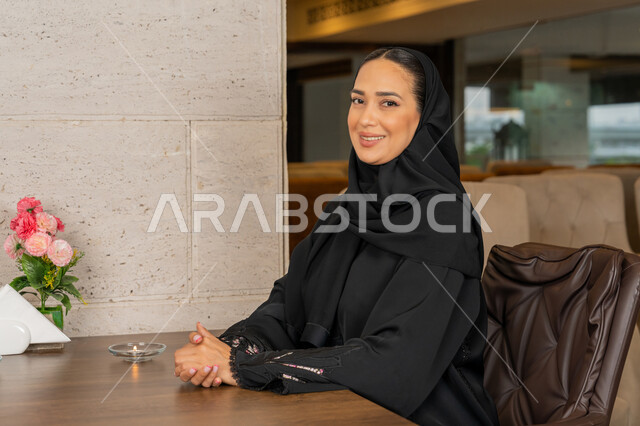 النظر إلى الكاميرا بإيماءات السعادة و السرور, الاستمتاع في أوقات الفراغ في إحدى المطاعم, قضاء الأوقات الممتعة في الخارج, امرأة عربية خليجية إماراتية محجبة مبتسمة ترتدي العباءة السوداء تجلس على مائدة الطعام في المطعم