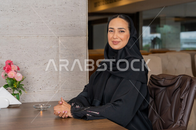 الاستمتاع في أوقات الفراغ في إحدى المطاعم, قضاء الأوقات الممتعة في الخارج, النظر إلى الكاميرا بإيماءات السعادة و السرور, امرأة عربية خليجية إماراتية محجبة مبتسمة ترتدي العباءة السوداء تجلس على مائدة الطعام في المطعم