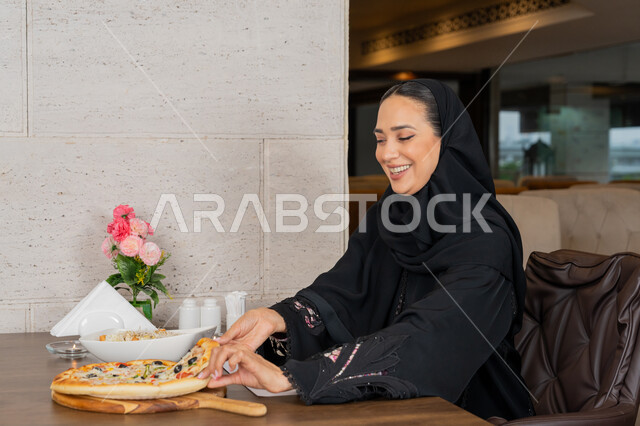 مطاعم و كافيهات في الإمارات, الاستمتاع بالأنشطة الترفيهية, قضاء الأوقات الممتعة في المطعم, امرأة عربية خليجية إماراتية محجبة ترتدي العباءة السوداء تجلس على المائدة و تقوم بتناول البيتزا, تشكيلة متنوعة و مختلفة من المأكولات