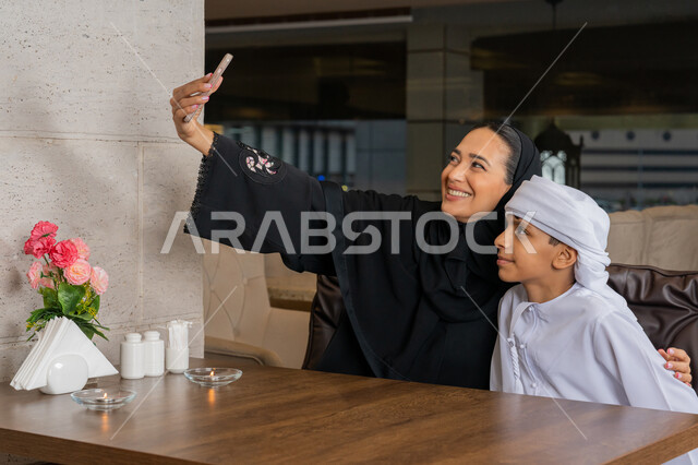 أنشطة ترفيهية مع الأسرة, توثيق اللحظات المميزة باستخدام الأجهزة التقنية الحديثة, قضاء الأوقات الممتعة مع الأبناء, امرأة عربية خليجية إماراتية مع ابنها يرتديان الزي التقليدي الإماراتي يقومان بالتقاط صور السيلفي التذكارية عبر الجوال, الأجواء العائلية السعيدة