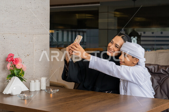 توثيق اللحظات المميزة باستخدام الأجهزة التقنية الحديثة, فعاليات و نشاطات مع الأسرة, قضاء الأوقات الممتعة مع الأبناء, امرأة عربية خليجية إماراتية مع ابنها يرتديان الزي التقليدي الإماراتي يقومان بالتقاط صور السيلفي التذكارية عبر الجوال, الأجواء العائلية السعيدة