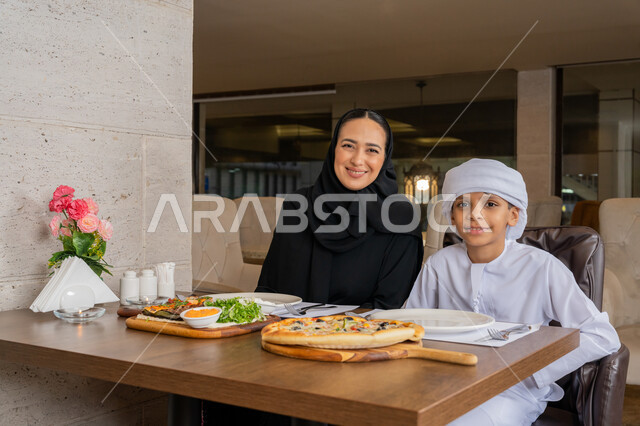 النظر إلى الكاميرا بإيماءات السعادة و السرور, أجواء عائلية سعيدة, قضاء الأوقات الممتعة مع الأبناء, امرأة عربية خليجية إماراتية محجبة ترتدي العباءة السوداء تجلس على مائدة الطعام مع ابنها و يقومان بتناول الغداء, مطاعم و كافيهات في الإمارات