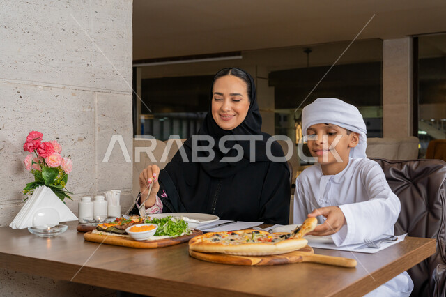 أجواء عائلية سعيدة, قضاء الأوقات الممتعة مع الأبناء, مفهوم الألفة و المحبة بين أفراد الأسرة, امرأة عربية خليجية إماراتية محجبة ترتدي العباءة السوداء تجلس على مائدة الطعام مع ابنها و يقومان بتناول الغداء, مطاعم و كافيهات في الإمارات
