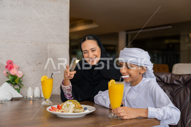 قضاء الأوقات الممتعة مع الأبناء, أجواء عائلية سعيدة, مفهوم الألفة و المحبة بين أفراد الأسرة, امرأة عربية خليجية إماراتية محجبة ترتدي العباءة السوداء تجلس على المائدة مع ابنها و تشير بإصبع السبابة إلى الطبق, مطاعم و كافيهات في الإمارات