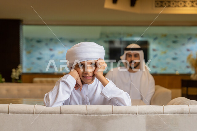 الأجواء الأسرية السعيدة, مشاركة الأبناء الأنشطة الترفيهية في الخارج, ولد عربي خليجي إماراتي يرتدي الكندورة و العمامة البيضاء يجلس مع والده في أحد المطاعم يضع يديه على خده بتعابير الملل و الضجر, قضاء الأوقات الممتعة مع العائلة