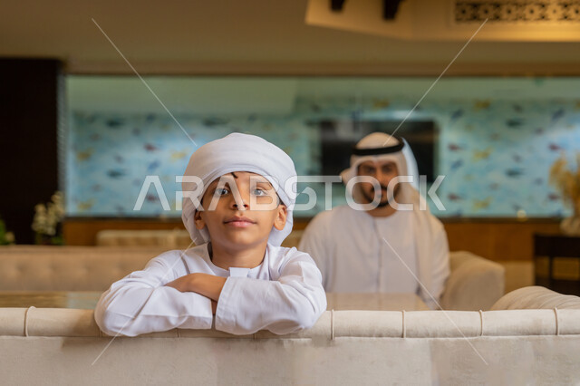 Looking at the camera with gestures of happiness and pleasure, spending quality time with family, an Emirati Gulf Arab man wearing a white kandura and ghutra sitting with his son in a restaurant, a happy family atmosphere, children participating in recreational activities outside