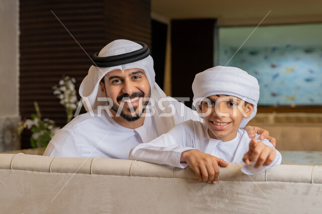 النظر إلى الكاميرا بإيماءات السعادة و السرور, الإشارة بإصبع السبابة إلى الأمام, رجل عربي خليجي إماراتي يرتدي الكندورة و الغترة البيضاء يجلس بجانب ابنه و يضع يده على كتفه بتعابير الدعم و المحبة, قضاء الأوقات الممتعة مع الأبناء, الأجواء العائلية السعيدة