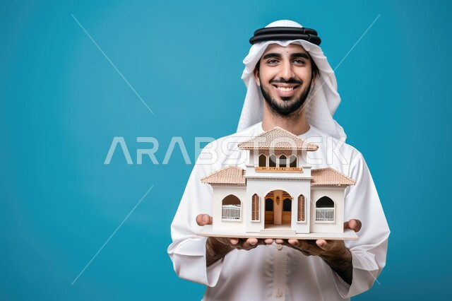 مفهوم الاسكان والتمليك، ايماءات تعبر عن السعادة بشراء عقار جديد, بورتريه مقرب لرجل عربي خليجي سعودي يرتدي الثوب التقليدي يحمل بيده مجسم بيت جديد، النظر الى الكاميرا بتعابير الفرح، امتلاك سكن حديث والاستقرار العائلي، خلفية زرقاء