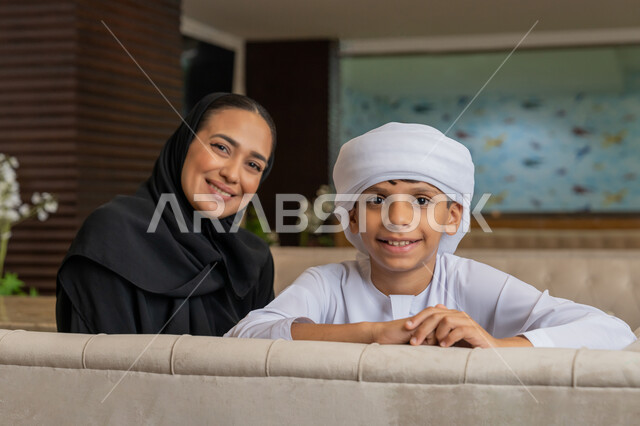 النظر إلى الكاميرا بإيماءات السعادة و السرور, مفهوم الألفة و المحبة بين أفراد الأسرة, أجواء عائلية سعيدة مليئة بالحنان, امرأة عربية خليجية إماراتية محجبة ترتدي العباءة السوداء تجلس بجانب ابنها في المطعم, قضاء الأوقات الممتعة مع الأبناء