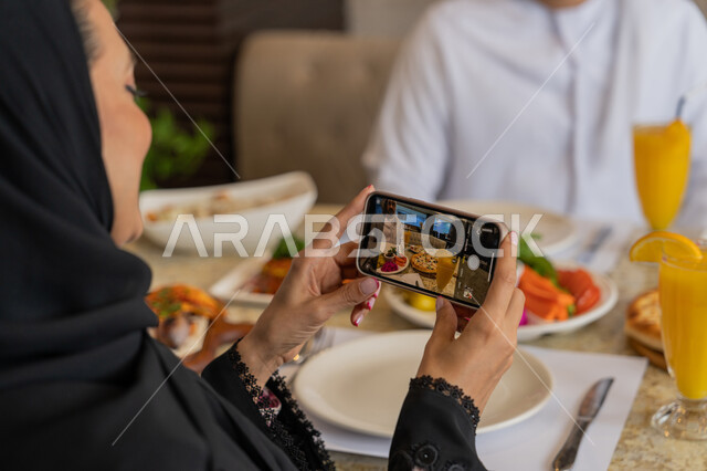 الاستمتاع بأجواء المطاعم في الإمارات, صورة مقربة لامرأة عربية خليجية إماراتية ترتدي الزي التقليدي الإماراتي تجلس مع زوجها في المطعم و تقوم بالتقاط الصور لمائدة الطعام عبر الجوال, تشكيلة مختلفة من الأطعمة على المائدة