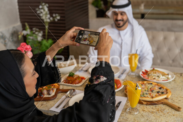 Taking souvenir photos using mobile phone, spending fun family time, an Arab Gulf Emirati couple wearing traditional Emirati dress having lunch, enjoying the atmosphere of restaurants in the Emirates, a variety of foods on the table