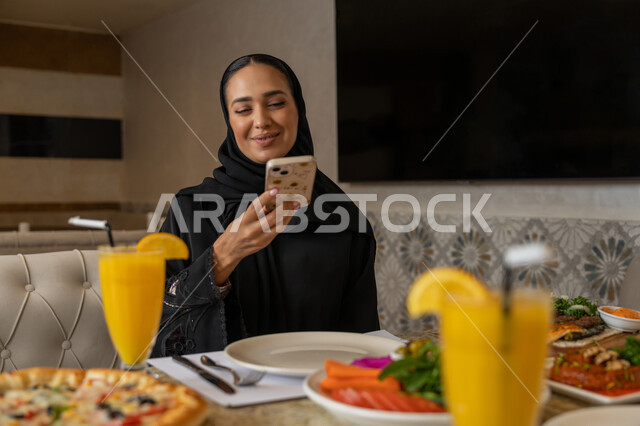 تشكيلة متنوعة و مختلفة من المأكولات والمشروبات, قضاء الأوقات الممتعة في المطعم, استخدام الأجهزة التقنية الحديثة, امرأة عربية خليجية إماراتية ترتدي الزي التقليدي الإماراتي تجلس على المائدة و تقوم بالتقاط الصور التذكارية عبر الجوال, مطاعم و كافيهات في الإمارات