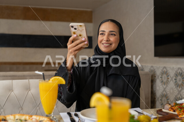 قضاء الأوقات الممتعة في المطعم, استخدام الأجهزة التقنية الحديثة, امرأة عربية خليجية إماراتية ترتدي الزي التقليدي الإماراتي تجلس على المائدة و تقوم بالتقاط الصور التذكارية عبر الجوال, تشكيلة متنوعة و مختلفة من المأكولات والمشروبات, مطاعم و كافيهات في الإمارات