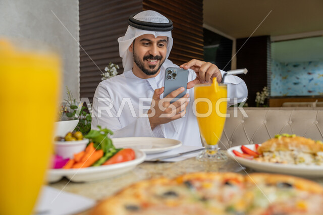 تشكيلة متنوعة و مختلفة من المأكولات والمشروبات, , قضاء الأوقات الممتعة في المطعم, رجل عربي خليجي إماراتي يرتدي الكندورة و الغترة البيضاء يجلس على مائدة الطعام و يقوم بالتقاط الصور التذكارية باستخدام الجوال, مطاعم و كافيهات في الإمارات
