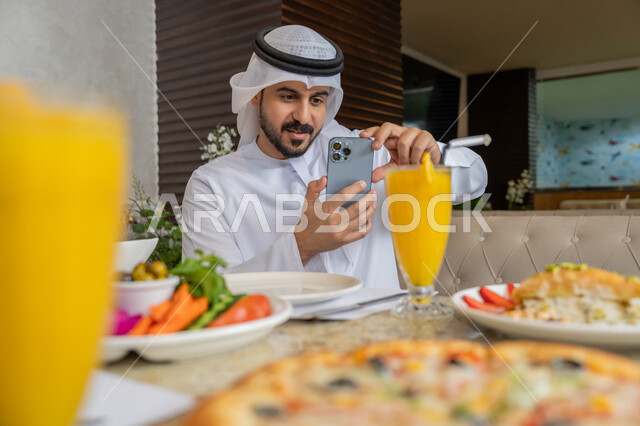مطاعم و كافيهات في الإمارات, قضاء الأوقات الممتعة في المطعم, رجل عربي خليجي إماراتي يرتدي الكندورة و الغترة البيضاء يجلس على مائدة الطعام و يقوم بالتقاط الصور التذكارية باستخدام الجوال, تشكيلة متنوعة و مختلفة من المأكولات والمشروبات