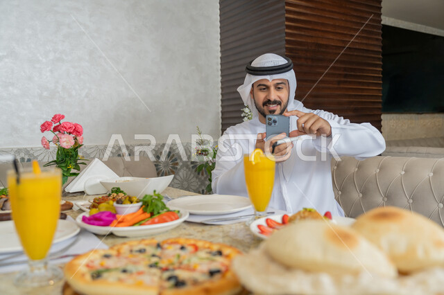 Having a good time in the restaurant, an Emirati Gulf Arab man wearing a white kandura and ghutra sitting at the dining table and taking souvenir photos using his mobile phone, a diverse and different selection of foods and drinks, restaurants and cafes in the Emirates
