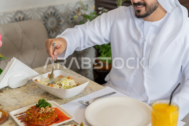الاستمتاع بتناول الأطباق الشهية, مطاعم و كافيهات في الإمارات, قضاء الأوقات الممتعة في المطعم, صورة مقربة لرجل عربي خليجي إماراتي يرتدي الكندورة و الغترة البيضاء يجلس على المائدة يقوم بتناول الغداء, تشكيلة متنوعة و مختلفة من المأكولات والمشروبات