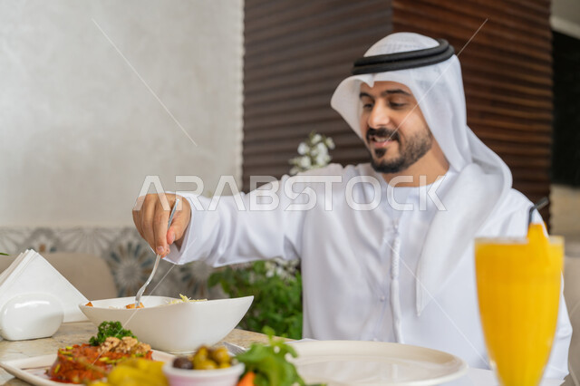 مطاعم و كافيهات في الإمارات, قضاء الأوقات الممتعة في المطعم, رجل عربي خليجي إماراتي يرتدي الكندورة و الغترة البيضاء يجلس على المائدة يقوم بتناول الغداء, أطباق متنوعة و مختلفة من المأكولات والمشروبات, الاستمتاع بتناول الأطباق الشهية