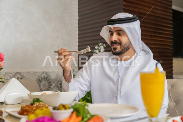 Enjoying delicious dishes, having a good time in the restaurant, an Emirati Gulf Arab man wearing a white kandura and ghutra sitting at the table eating lunch, a diverse and different selection of foods and drinks, restaurants and cafes in the Emirates