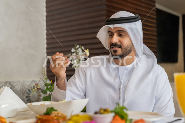 قضاء الأوقات الممتعة في المطعم, رجل عربي خليجي إماراتي يرتدي الكندورة و الغترة البيضاء يجلس على المائدة يقوم بتناول الغداء, تشكيلة متنوعة و مختلفة من المأكولات والمشروبات, مطاعم و كافيهات في الإمارات, الاستمتاع بتناول الأطباق الشهية