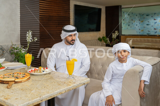 قضاء الأوقات العائلية الممتعة, رجل عربي خليجي إماراتي يرتدي الكندورة و الغترة البيضاء يجلس مع ابنه على مائدة الطعام, مطاعم و كافيهات في الإمارات, تشكيلة متنوعة من المأكولات على سفرة الأكل, وصفات و أطباق شهية