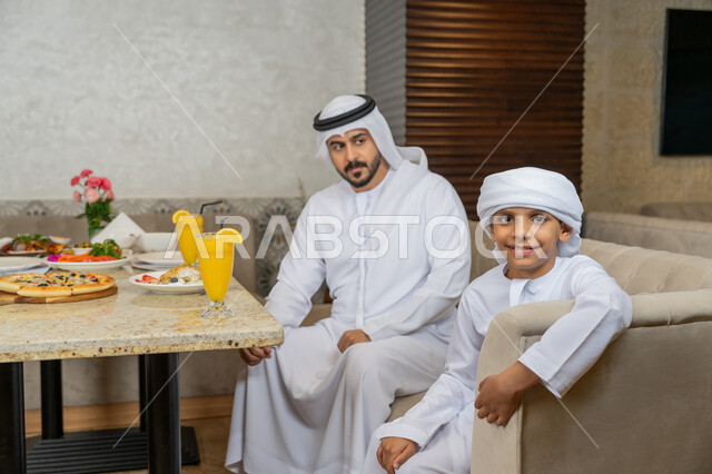 قضاء الأوقات العائلية الممتعة, رجل عربي خليجي إماراتي يرتدي الكندورة و الغترة البيضاء يجلس مع ابنه على مائدة الطعام, مطاعم و كافيهات في الإمارات, تشكيلة متنوعة من المأكولات على سفرة الأكل, وصفات و أطباق شهية