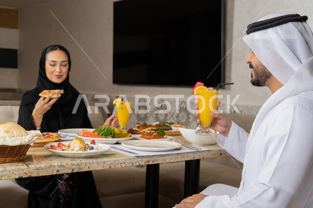 قضاء الأوقات العائلية الممتعة, تشكيلة مختلفة من الأطعمة على المائدة, الاستمتاع بأجواء المطاعم في الإمارات, زوجان عربيان خليجيان إماراتيان يرتديان الثوب التقليدي الإماراتي يقومان بتناول الغداء, أصناف متنوعة من المأكولات الشهية