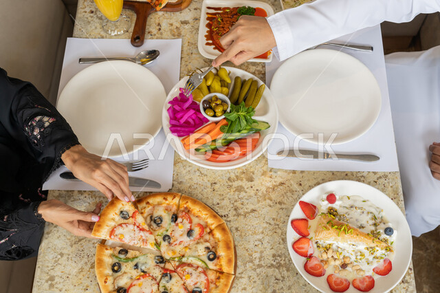 قضاء أوقات رائعة في المطعم, صورة من الأعلى لمائدة طعام لزوجان عربيان خليجيان إماراتيان يرتديان الثوب التقليدي الإماراتي يقومان بتناول الغداء، تشكيلة متنوعة من المأكولات الشهية على السفرة، أجواء عائلية فريدة و مميزة، مطاعم و كافيهات في الإمارات