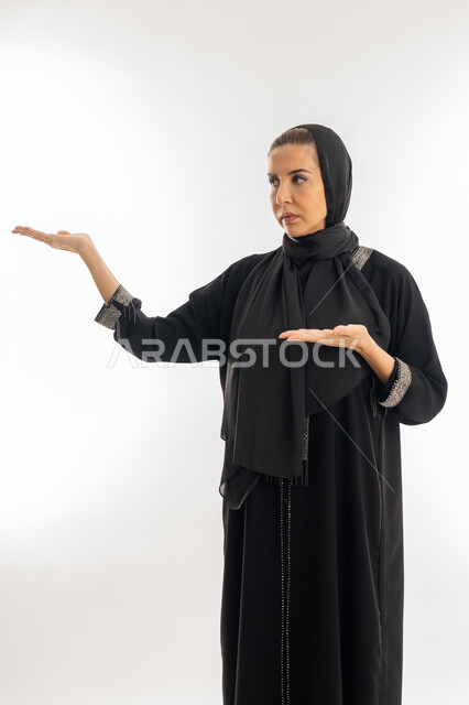 إشارات وحركات يدين تدل على هذا المكان (هنا)، سيدة سعودية تقوم بتحديد موقع شيء معين، بورتريه لامرأة عربية خليجية إماراتية ترتدي عباءة وحجاب تشير بيديها إلى جهة اليسار، خلفية بيضاء