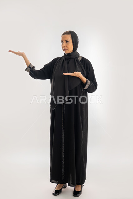 إشارات وحركات يدين تدل على هذا المكان (هنا)، سيدة سعودية تقوم بتحديد موقع شيء معين، بورتريه لامرأة عربية خليجية إماراتية ترتدي عباءة وحجاب تشير بيديها إلى جهة اليسار، صورة الجسم بالطول الكامل، خلفية بيضاء