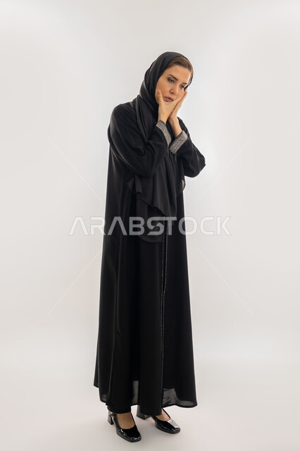 الشعور بألم الأسنان وتسوس الأضراس, سيدة سعودية بحاجه لزيارة طبيب الأسنان، بورتريه لإمراة عربية إماراتية خليجية ترتدي العباءة والحجاب تضع يدها على خدها، إيماءات وجه ويدين تدل على الالم ووجع الفم، الجسم بالطول الكامل، خلفية بيضاء