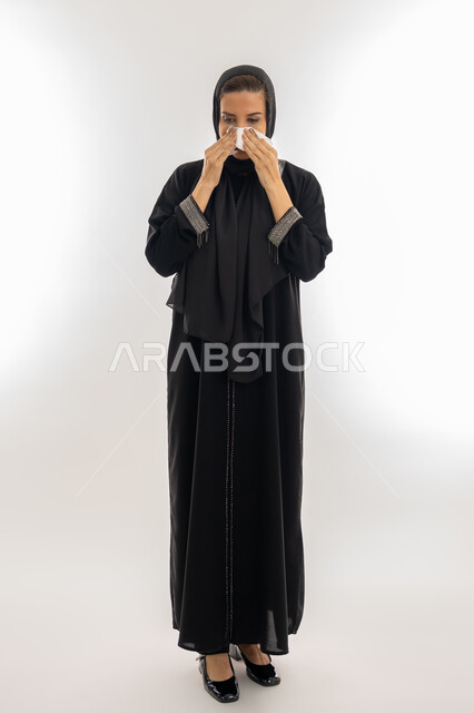 استعمال المناديل الورقيه أثناء العطس، الإصابة بالزكام والانفلونزا, سيدة سعودية تقف بتعابير تدل على العطاس والإرهاق، الشعور بالتعب والمرض، بورتريه لإمرأة عربية إماراتية خليجية محجبة ترتدي العباءة تعاني من الحساسية, الجسم بالطول الكامل, خلفية بيضاء
