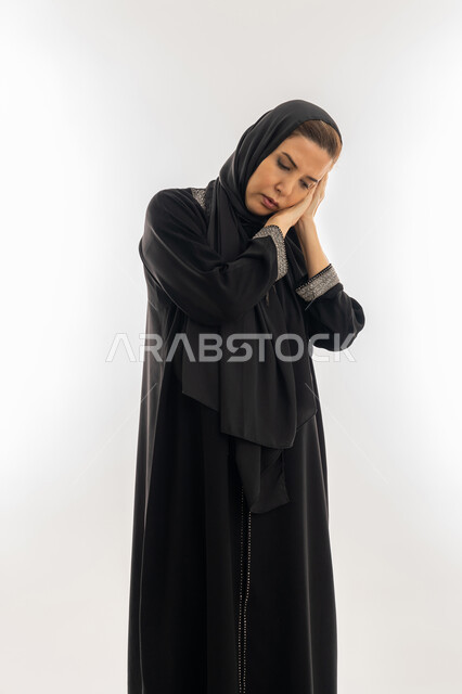 السهر لوقت متأخر من الليل، سيدة سعودية تشعر بالكسل والخمول و الارهاق، الحاجة للنوم والراحة، بورتريه  لإمرأه عربية خليجية اماراتية محجبة ترتدي العباءة السوداء تضع يديها تحت رأسها بإيماءات النعاس والتعب، خلفية بيضاء