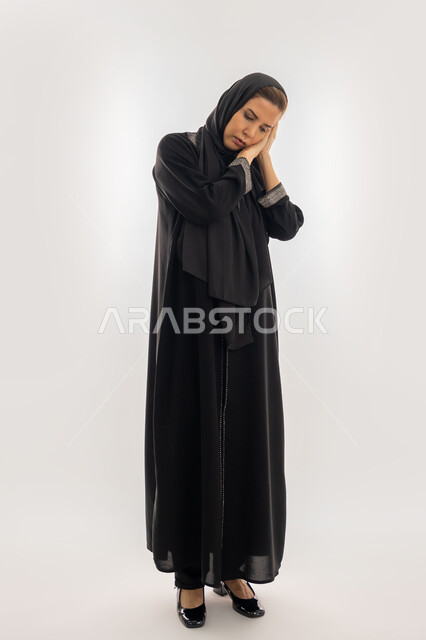 السهر لوقت متأخر من الليل، سيدة سعودية تشعر بالكسل والخمول و الارهاق، الحاجة للنوم والراحة، بورتريه  لإمرأه عربية خليجية اماراتية محجبة ترتدي العباءة السوداء تضع يديها تحت رأسها بإيماءات النعاس والتعب، صورة الجسم بالطول الكامل، خلفية بيضاء