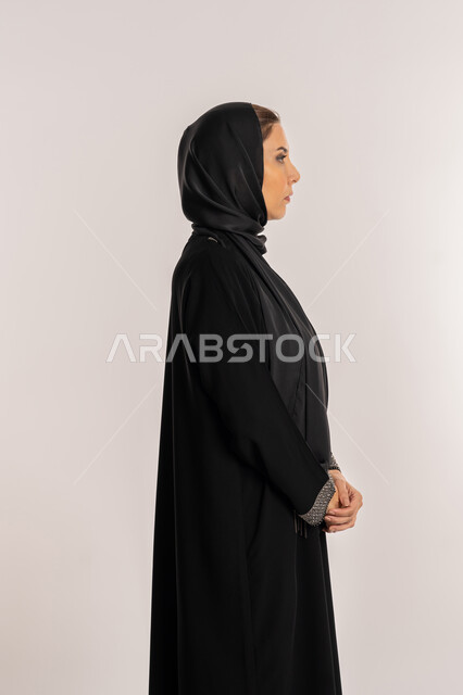 مفهوم الاناقة و الاهتمام بالمظهر الخارجي و الأنوثة, بورتريه من الجانب لامرأة عربيه إماراتية محجبة ترتدي العباءه السوداء تقف بأيدي متشابكة, سيدة سعودية مبتسمة تقف باستقامة بايماءات الثقة بالنفس, خلفية بيضاء
