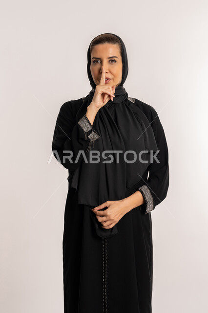 التوقف عن الكلام و الأمر بالصمت و الاستماع، بورتريه لإمرأة عربية إماراتية خليجية محجبة ترتدي العباءة والحجاب تنظر إلى الكاميرا وترفع اصبعها الى فمها، سيدة سعودية تقف بإيماءات طلب الهدوء وعدم الإزعاج, خلفية بيضاء