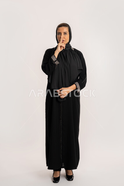 التوقف عن الكلام و الأمر بالصمت و الاستماع، بورتريه لإمرأة عربية إماراتية خليجية محجبة ترتدي العباءة والحجاب تنظر إلى الكاميرا وترفع اصبعها الى فمها، سيدة سعودية تقف بإيماءات طلب الهدوء وعدم الإزعاج, صورة الجسم بالطول الكامل، خلفية بيضاء