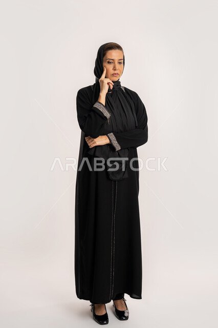التمعن والتركيز والتخطيط للمستقبل، سيدة سعودية تقوم بمحاولة إيجاد حل لمشكلة ما، بورتريه لإمرأة عربية خليجية إماراتية محجبة ترتدي عباءة وردية تضع يدها على ذقنها وتنظر بإيماءات التفكير والتأمل، صورة الجسم بالطول الكامل، خلفية بيضاء