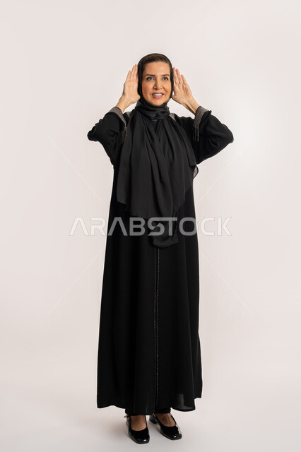 سماع الأحاديث و النقاشات، محاولة التجسس والتنصت, بورترية لإمراة عربية إماراتية خليجية ترتدي العباءة والحجاب تضع يديها خلف اذنيها، سيدة سعودية تقف بإيماءات تدل على الإنصات وكشف الاسرار، صورة الجسم بالطول الكامل، خلفية بيضاء